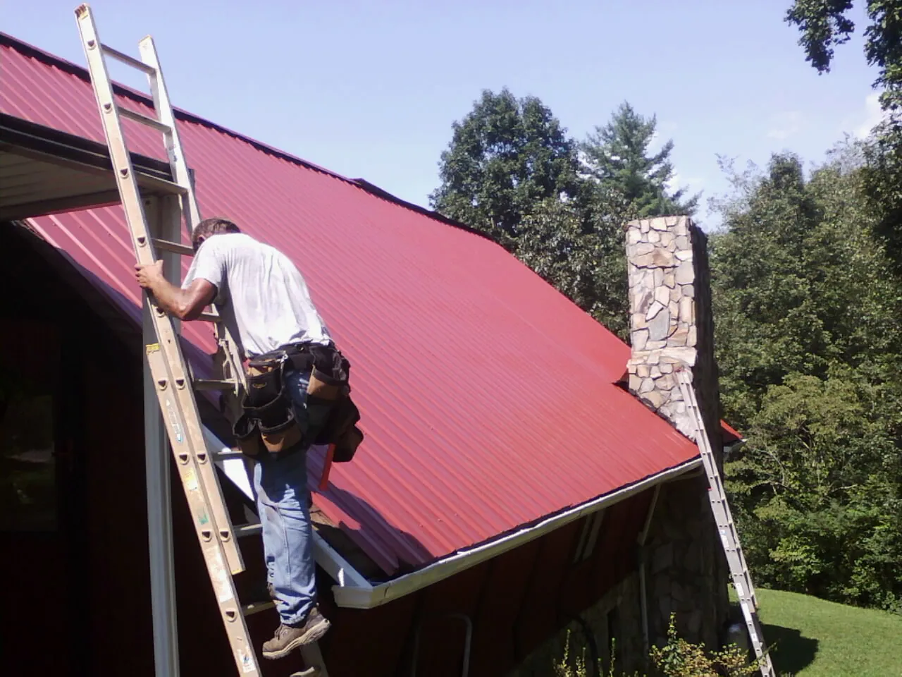 Roofer on a ladder beside a red metal roof and stone chimney — Soffit & Fascia Repair by Roofing Brothers in Loves Park
